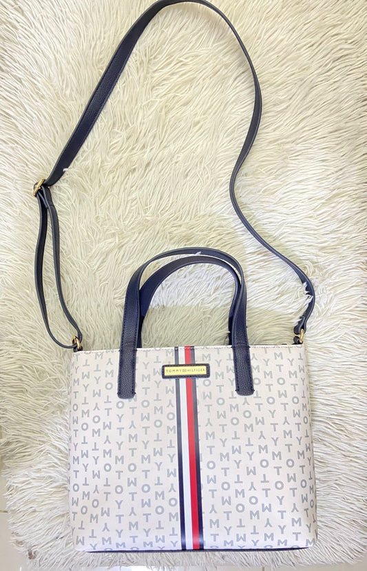 Cartera Tommy Hilfiger Original, color blanco con estampado de la marca, placa de la marca al frente en color dorado, linea en el frente color rojo, blanco y azul marino, correa adicional delgada.