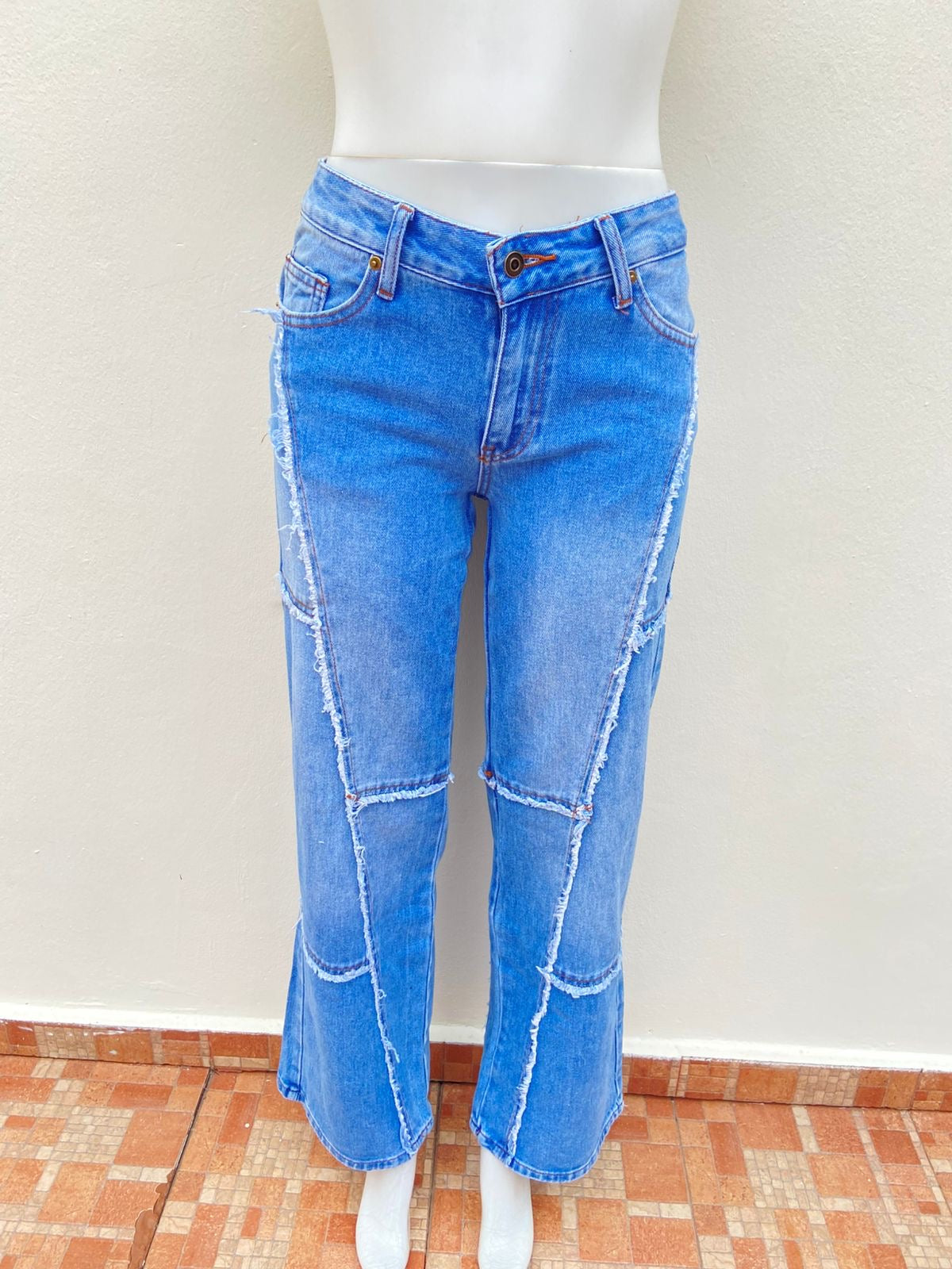 Pantalón Jean, azul claro con costura en hilo marrón y vuelos del Jean destruido en forma de líneas.